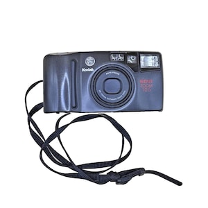 Puede incluir: Cámara Kodak Star Zoom 105 negra con correa negra. La cámara tiene una lente, flash y las palabras "Kodak" y "Star Zoom 105" impresas. La correa está sujeta a la cámara.