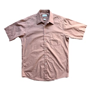 Puede incluir: Camisa de manga corta en color rosa apagado. Presenta un cuello clásico, un bolsillo en el pecho y una botonadura frontal. La tela parece ser una mezcla ligera de algodón o lino, adecuada para climas cálidos.