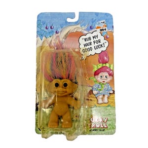 Può includere: Una bambola Lucky Troll nella sua confezione originale. La bambola ha un corpo abbronzato, grandi occhi e capelli colorati e appuntiti. La confezione presenta un'illustrazione di un troll e il testo "RUB MY HAIR FOR GOOD LUCK!"