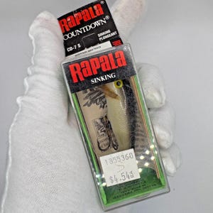 May include: A Rapala Countdown CD-7 S sinking fishing lure in a clear plastic package. The lure is silver, black, and cream-colored, with a yellow eye. The packaging includes the Rapala logo and product information.