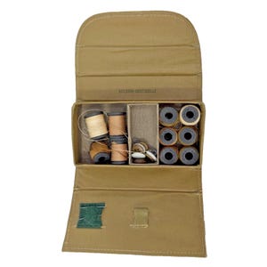 Könnte beinhalten: Ein beigefarbenes Nähset-Etui aus Stoff mit der Aufschrift BELDING-CORTICELLI. Das geöffnete Etui enthält mehrere Garnrollen in verschiedenen Braun- und Beigetönen, ein kleines Fach mit verschiedenen Knöpfen und ein Nadelkissen aus grünem Filz.