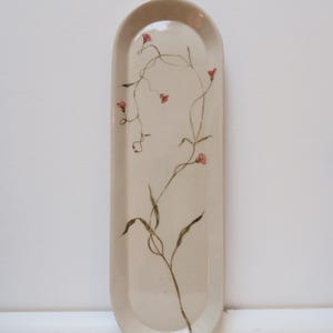 May include: A cream-coloured, oval ceramic tray with a hand-painted floral design. The design features delicate brown stems, green leaves, and small red flowers. The tray has rounded edges and a smooth surface.