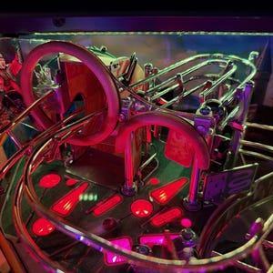 May include: A pinball machine with a fantasy theme. The playfield features a complex arrangement of ramps, bumpers, and targets, illuminated by red and purple lights. The backglass depicts a castle scene. The machine's design incorporates metallic and transparent elements.