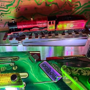 Puede incluir: Un tren de juguete rojo y negro sobre una vía plateada, parte de una máquina de pinball. La máquina de pinball tiene detalles en verde y amarillo, con la palabra "PROTEGO!" en un letrero morado. El fondo es una mezcla de verde y marrón.