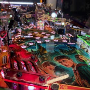 May include: A colorful pinball machine featuring a Harry Potter theme. The playfield showcases images of the characters and scenes from the movies. The machine has red, green, and blue lighting, with various ramps, bumpers, and targets.