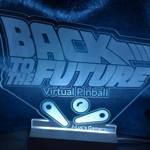 May include: A white acrylic sign with a blue glow, featuring the text "Back to the Future Virtual Pinball" and a stylized image of a pinball machine with flippers and a ball. The sign also includes the text "J-Luc's Gameroom".