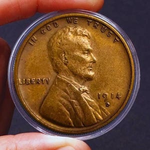 May include: A copper-coloured 1914 Lincoln penny with the words "In God We Trust" and "Liberty" inscribed on the coin. The coin is in a clear plastic coin holder.