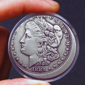 May include: A silver 1884 Morgan dollar coin in a protective case. The coin features a profile of Liberty and the inscription "LIBERTY." The reverse side of the coin has the words "E PLURIBUS UNUM" and "UNITED STATES OF AMERICA."