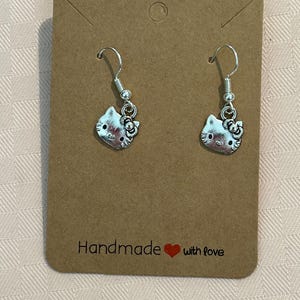 May include: Silver-coloured Hello Kitty earrings hanging from hooks. The earrings are displayed on a brown card with the text "Handmade with love". The background is a light pink patterned fabric.