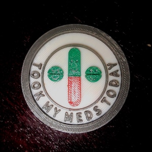 May include: A circular, white and grey 3D-printed object with the text "TOOK MY MEDS TODAY". The centre shows a green and red pill with a smiling face, with two green smiling pill-shaped designs. This item is likely a reminder or a novelty item.