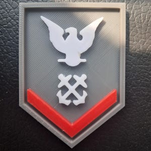 Op de afbeelding: Een grijs en rood embleem met een witte adelaar bovenaan en een wit ankerontwerp eronder. Onderaan staat een rode chevron. Het embleem is een 3D-geprint ontwerp.