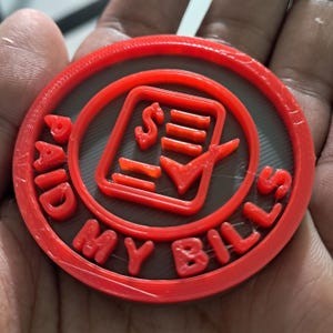 Puede incluir: Un token rojo, circular, impreso en 3D con las palabras "PAID MY BILLS" en letras en relieve alrededor del borde. El centro presenta una marca de verificación, un signo de dólar y líneas, todo en rojo, sobre un fondo gris.