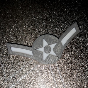 May include: A grey and white 3D-printed emblem. It features a star inside a circle and two white bars. The item has a textured surface and is likely a decorative or symbolic item.