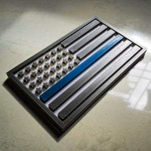 May include: A rectangular, gray and blue flag design with a blue stripe, gray stripes, and gray stars. The flag is set in a gray frame. The design is a 3D-printed item.