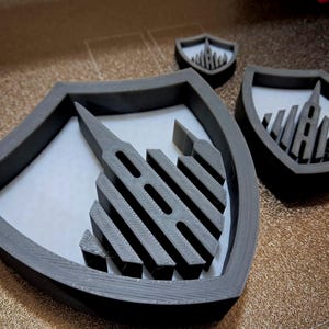 Könnte beinhalten: Graue, 3D-gedruckte Ausstecher in Schildform mit einem stilisierten architektonischen Design. Das Set besteht aus drei Ausstechern in verschiedenen Größen, jeweils mit detailliertem Innendesign und einem stabilen Rand. Die Ausstecher liegen auf einer hellen Oberfläche.