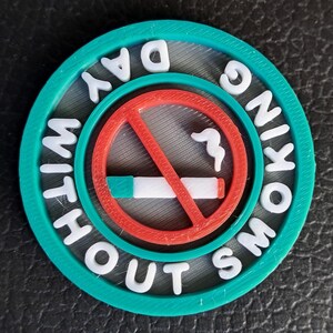 May include: A round, teal and gray emblem with the words "WITHOUT SMOKING DAY" in white, surrounding a red circle with a no-smoking symbol. The center features a cigarette with smoke.