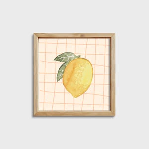 May include: A framed watercolor painting of a yellow lemon with green leaves on a peach background with a grid pattern.