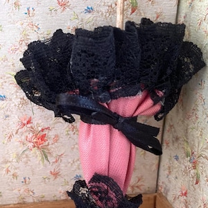 May include: A miniature pink parasol with black lace trim and a black bow. The parasol has a wooden handle and a rounded top. The lace trim is detailed and adds a delicate touch to the design.