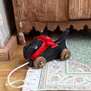 May include: A miniature black wooden dog pull toy with a red ribbon around its neck. The toy has small wooden wheels and a white string for pulling. The toy is on a patterned rug.