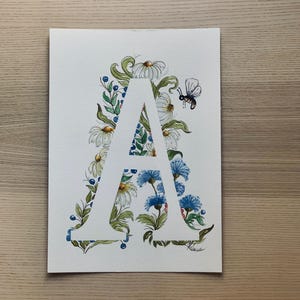 Letra floral en acuarela: Letra pintada a mano con acuarela, flores y bayas azules, ideal para decorar la habitación de un bebé o una boda.