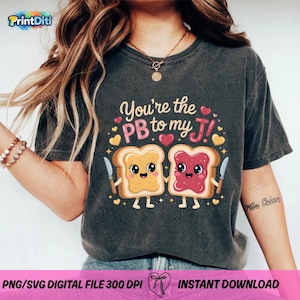 以下が含まれることがあります： 「You're the PB to my J!」の文字と、ピーナッツバターとジャムを塗った2枚のパンのグラフィックが描かれた、濃いグレーのTシャツ。デザインにはハートとナイフが含まれています。