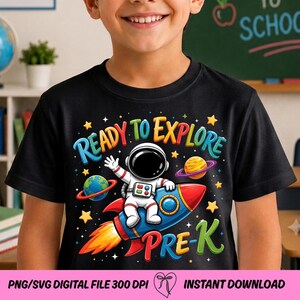 May include: A black t-shirt featuring a colorful graphic of an astronaut riding a blue and red rocket. The text reads "READY TO EXPLORE" and "PRE-K" in vibrant, multi-colored letters. The design includes yellow stars and two planets.