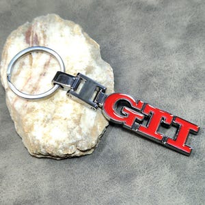 以下が含まれることがあります： 赤い「GTI」エンブレムが付いたシルバーカラーのメタルキーホルダー。キーホルダーにはスプリットリングと長方形の留め金が付いています。エンブレムは赤色で、シルバーの縁取りがあります。キーホルダーは明るい色の岩の上に置かれています。