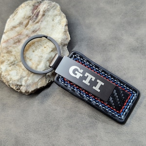 Peut inclure: Un porte-clés noir et argent avec les lettres "GTI" en blanc. Le porte-clés a une bordure en cuir noir avec des coutures bleues, rouges et blanches. Un porte-clés argenté est attaché au porte-clés.