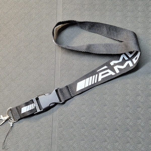 Könnte beinhalten: Schwarzes Schlüsselband mit weißem "AMG"-Text und Logo. Das Schlüsselband hat eine schwarze Kunststoffschnalle und einen silberfarbenen Metallclip zum Befestigen von Schlüsseln oder einem Ausweis. Das Schlüsselband ist aus gewebtem Stoff.