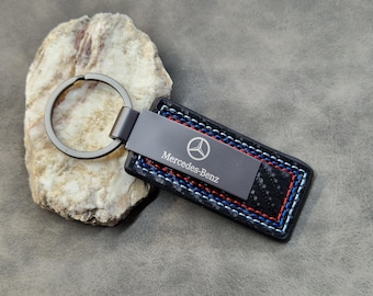Może przedstawiać: Czarny i srebrny brelok do kluczy Mercedes-Benz z wzorem z włókna węglowego i czerwonymi, białymi i niebieskimi szwami. Brelok ma metalowy pierścień, a logo i tekst Mercedes-Benz są widoczne.