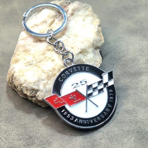 May include: A silver metal Corvette 25th Anniversary keyring. The round emblem has the words "CORVETTE" and "ANNIVERSARY 1978" around a white centre with the number "25" and crossed checkered flags.