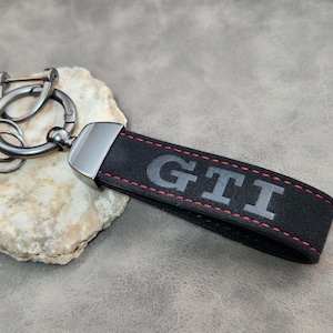 Peut inclure: Porte-clés en cuir noir avec les lettres "GTI" en gris, cousues avec du fil rouge. Le porte-clés est doté d'un fermoir et d'anneaux en métal argenté. Le fond est gris neutre.