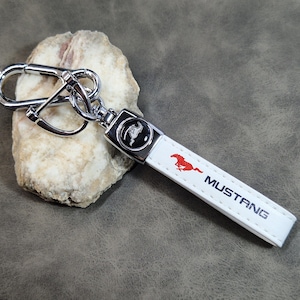 Puede incluir: Llavero de cuero blanco con mosquetón plateado y logotipo Mustang rojo. La palabra "MUSTANG" está impresa en azul. El llavero está sobre una piedra.