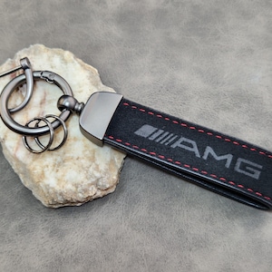 Könnte beinhalten: Schwarzer Schlüsselanhänger mit den Buchstaben "AMG" auf einem schwarzen Riemen mit roten Nähten. Der Schlüsselanhänger hat einen Metallverschluss und Schlüsselringe. Der Hintergrund ist eine neutrale graue Oberfläche.