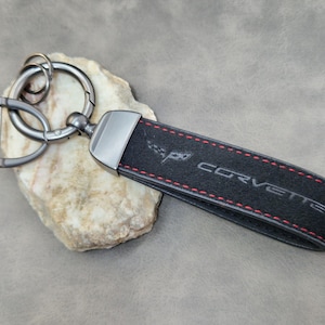 May include: Black key fob with the word "CORVETTE" and a logo stitched in red thread. The key fob is attached to a silver key ring and a silver clasp. The key fob is resting on a light-coloured rock.
