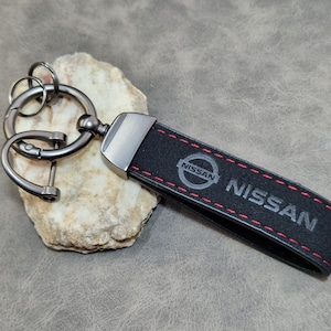 Può includere: Portachiavi in pelle nera con logo e testo Nissan, cuciture rosse. Ha una chiusura e un anello in metallo argentato. Lo sfondo è una superficie grigia neutra.