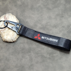 Könnte beinhalten: Schwarzes Mitsubishi Schlüsselband mit Metallverschluss und Schlüsselring. Das Band zeigt das Mitsubishi-Logo in Rot und den Markennamen in Weiß. Für Autoschlüssel.