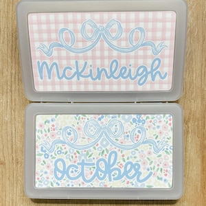 May include: Two rectangular, gray plastic cases. One case has a pink and white gingham pattern with the name "McKinleigh" in light blue script. The other case has a floral pattern with the word "October" in light blue script.