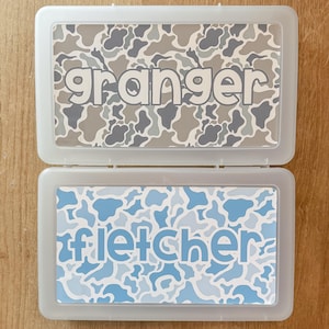 May include: Two rectangular, translucent plastic pencil cases stacked vertically. The top case features a gray and white camouflage pattern with the name granger printed in lowercase letters. The bottom case has a blue and white camouflage pattern with the name fletcher printed in lowercase letters.