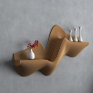 Op de afbeelding: Een modern, golfvormig houten rek aan een grijze muur. Het rek bevat witte vazen en een kleine vaas met rode bessen. Rechts staat een zwarte vaas met tarwearen.