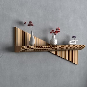 Op de afbeelding: Een modern houten wandrek met een uniek geometrisch ontwerp. Het rek bevat drie witte vazen met rode en paarse bloemen. Rechts staat een zwarte vaas met tarwearen. De achtergrond is een grijze muur.