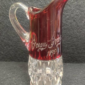 May include: A small, clear glass pitcher with a ruby red upper section and a clear, diamond-cut base. The words "Ocean Grove 1895" are etched in white script on the red portion. The pitcher has a clear, curved handle.