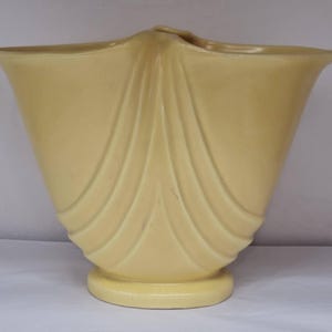 May include: A pale yellow ceramic vase with a wide, flared opening and a fluted design. The vase has a rounded base and a smooth, glossy finish.