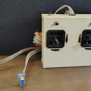 May include: A rectangular off-white control box featuring two black joystick assemblies, two small push buttons on the top face, and a single toggle switch. Multi-colored ribbon cables extend from the unit, terminating in two blue 9-pin connectors.