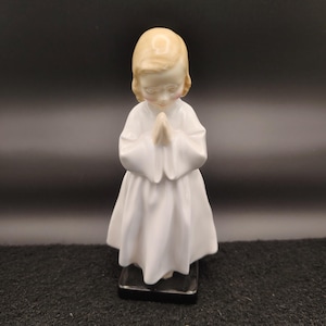 May include: A porcelain figurine of a young person in a white robe, with hands clasped in prayer. The figurine has blonde hair and a serene expression. It stands on a black rectangular base, set against a dark background.
