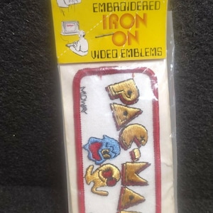 May include: Embroidered iron-on video game emblem featuring the Pac-Man logo. The patch has a red border and includes the text "MIDWAY" and "©88". The packaging is yellow with illustrations and the words "EMBROIDERED IRON ON VIDEO EMBLEMS".