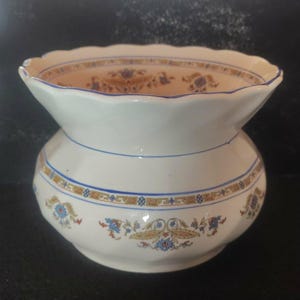 May include: A white ceramic vase with a flared, scalloped rim. The vase features blue and gold floral patterns and a blue band around the body. The interior of the rim is a light tan color.
