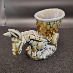 May include: A ceramic donkey figurine with a basket on its back. The donkey is light brown with darker brown accents, and the basket is a woven design. The figurine has a glossy finish and is likely a decorative item.