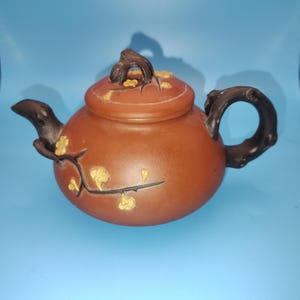 May include: A round, reddish-brown clay teapot featuring a dark brown handle and spout shaped like tree branches. The body and lid are decorated with raised, dark brown branch motifs accented with small, pale yellow flower blossoms.