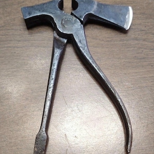 May include: A vintage, multi-tool metal tool with a hammer head, axe head, and screwdriver end. The tool is dark gray and appears to be made of steel. The tool is designed with a hinged mechanism and curved handles.